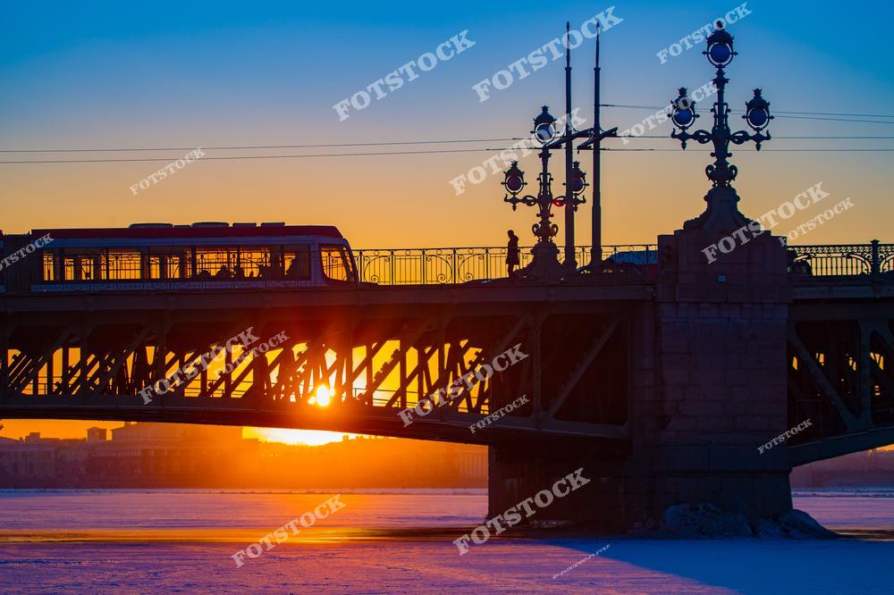 На фотографии запечатлен Троицкий мост в Санкт-Петербурге в момент захода солнца. Силуэт трамвая, пересекающего мост, выглядит эффектно на фоне золотого закатного неба. Замерзшая Нева и теплое свечение солнца создают контраст зимнего пейзажа и тепла света. Этот мост является одним из знаковых мест города, соединяя исторический центр с Петроградской стороной.